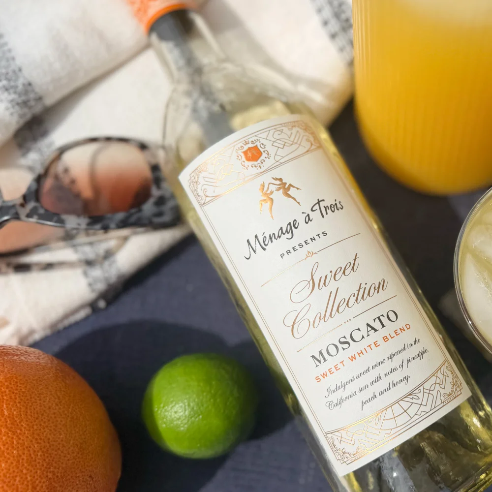 Bottle of Ménage à Trois Light Moscato Sweet White Blend white desert wine resting on a table cloth next to an orange and lime, sunglasses, orange juice.